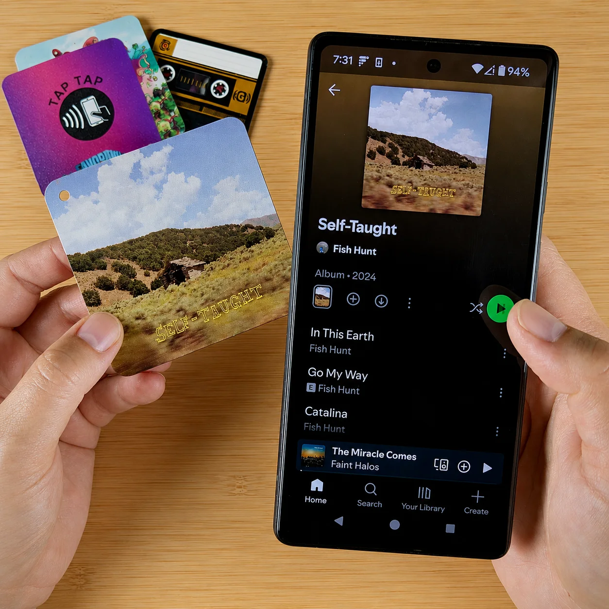 Hands holding a Fish Hunt 'Self-Taught' Custom Goodz card next to a phone showing the same album on Spotify, with a small stack of other Custom Goodz cards on the table.