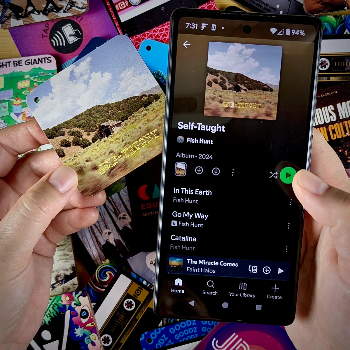 A hand holding a Custom Goodz card next to a phone playing the album it links to.