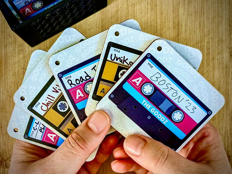 A fan of cassette-themed Mixtape Goodz cards held in two hands, with a small wooden crate of more Goodz in the background.