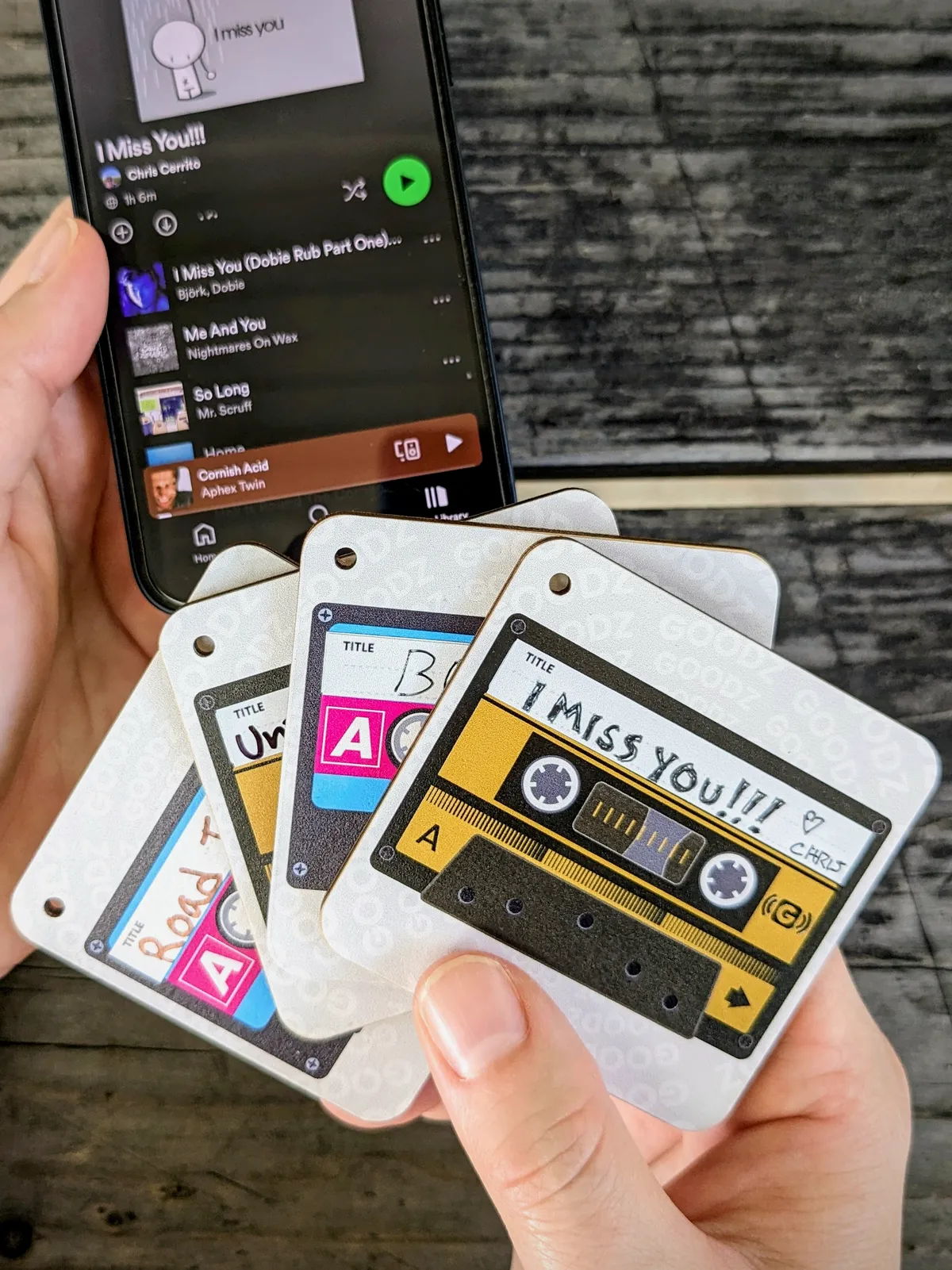 A hand holding a fan of cassette-themed Mixtape Goodz cards in front of a phone showing a Spotify playlist titled 'I Miss You!!!'.
